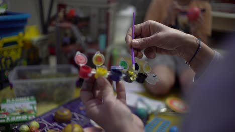 Cerca-De-Las-Manos-De-Una-Mujer,-Mezclando-Colores-De-Pintura-Con-Un-Pincel-Pequeño,-Su-Hijo-Pintando-Un-Planeta-De-Juguete-En-El-Fondo-Borroso-De-Su-Casa-4k