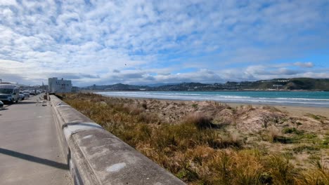 Scenic-view-of-Lyall-Bay-beach,-surf-waves-rolling-in-and-coastal-sand-dunes-on-a-blue-sky-sunny-day-in-capital-city-of-Wellington,-New-Zealand-Aotearoa