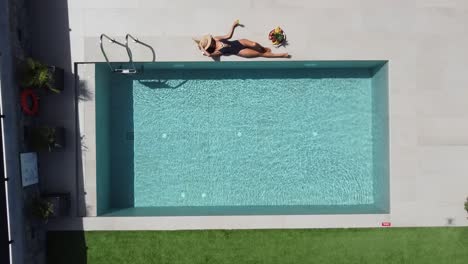 Mujer-Delgada-De-Cabello-Oscuro-Tumbada-Al-Borde-De-Una-Hermosa-Piscina-Al-Aire-Libre:-Viste-Un-Traje-De-Baño-Oscuro-Y-Disfruta-Del-Sol