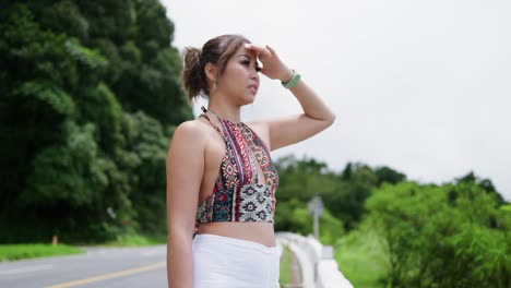 Woman-with-light-brown-hair-and-a-colorful-top-stands-by-the-roadside,-shielding-her-eyes-from-the-sun-as-she-looks-into-the-distance