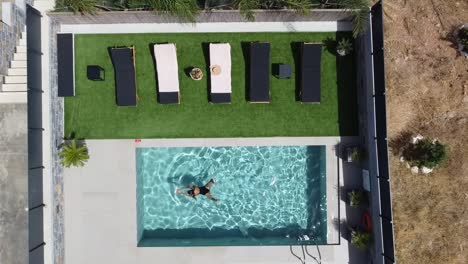 Dark-haired-slim-young-lady-is-enjoying-a-swim-in-a-nice-outdoor-pool---she-wears-a-dark-swimming-costume-and-enjoys-the-sun---drone-films-her-from-a-bird's-eye-view