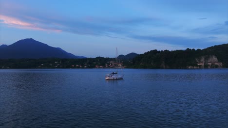 Luftaufnahme-Eines-Segelboots,-Das-In-Der-Abenddämmerung-Ruhig-Auf-Dem-Wasser-Vor-Anker-Liegt,-Mit-Majestätischen-Bergen-Im-Hintergrund-Und-Einem-Heiteren-Himmel-In-Blau--Und-Rosatönen