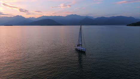 Un-Velero-Navega-Pacíficamente-En-Un-Lago-Tranquilo-Bajo-Un-Colorido-Cielo-Al-Atardecer,-Con-Una-Pintoresca-Cadena-Montañosa-Al-Fondo,-Capturando-Un-Momento-Sereno-Y-Relajante