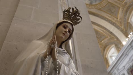 Estatua-De-La-Virgen-María-Adornada-Con-Una-Corona-Dorada-Y-Un-Rosario,-Con-El-Telón-De-Fondo-Del-Interior-De-Una-Iglesia-Ornamentada