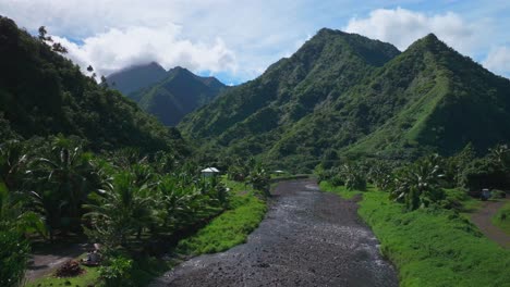 River-crossing-towering-mountain-volcano-peaks-Teahupoo-town-Tahiti-French-Polynesia-Moorea-Papeete-aerial-drone-stunning-island-late-morning-afternoon-blue-sky-daytime-sunny-clouds-upwards-motion