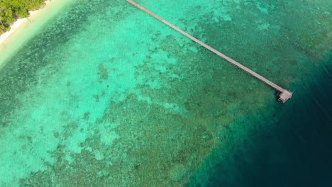 Eine-Luftaufnahme-Zeigt-Eine-Unberührte-Tropische-Küste-Mit-Einem-Langen-Pier,-Der-Sich-Bis-Ins-Türkisfarbene-Wasser-Der-Insel-Kri,-Raja-Ampat-In-Indonesien-Erstreckt.