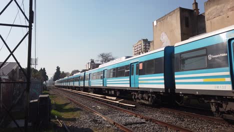 Un-Tren-Azul-Viaja-En-La-Línea-Sarmiento,-Servicio-De-Trenes-De-Cercanías-En-La-Provincia-De-Buenos-Aires,-Argentina,-Horizonte-Diurno-Y-Edificios-De-La-Ciudad-Alrededor-De-La-Zona-Verde