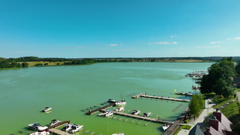 Aerial-view-of-the-marina-in-Ryn,-Warmia-Masuria,-with-boats-docked-in-a-green-lake-and-expansive-countryside-in-the-background,-capturing-the-beauty-of-the-region