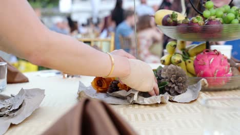 Mujer-En-Primer-Plano-Colocando-Flores-Y-Una-Piña-Para-Un-Centro-De-Mesa-De-Recepción-De-Boda-Con-Una-Bandeja-Escalonada-De-Frutas-Que-Contiene-Pitahaya-Roja,-Plátanos,-Mangostán-Y-Uvas