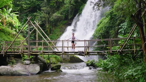 Standing-on-a-rustic-bamboo-bridge,-a-woman-enjoys-the-sight-of-a-powerful-waterfall-behind-her