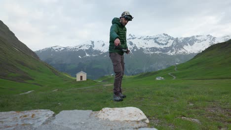 A-drone-pilot-with-goggles-stands-in-Swiss-mountains-and-catches-returning-drone-in-hand