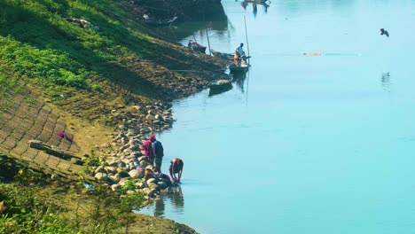 Villagers-in-a-rural-area-of-Bangladesh,-washing-clothes-on-riverbank-while-a-fishing-boat-pulls-in-a-fishing-net