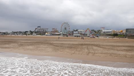 Una-Amplia-Playa-De-Arena-En-Skegness-Se-Extiende-Hacia-Un-Parque-De-Diversiones-Con-Una-Noria,-Montañas-Rusas-Y-Varias-Atracciones-Al-Fondo.