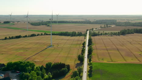 Vista-Aérea-De-Un-Largo-Camino-Flanqueado-Por-Campos-De-Trigo-Dorado-Y-Turbinas-Eólicas-En-La-Distancia,-Capturando-La-Vasta-Extensión-Rural