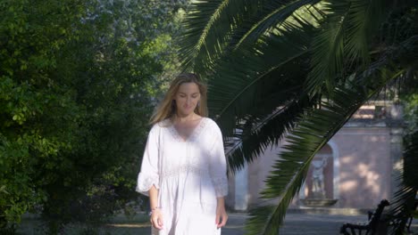 A-joyful-young-blonde-woman-in-a-white-dress-is-walking-in-the-summer-park