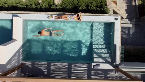 Brünette-Frau-Im-Badeanzug-Liegt-Am-Beckenrand-Im-Cayo-Resort-Auf-Kreta,-Griechenland---Mann-Schwimmt-Und-Taucht-Im-Pool---Drohne-Fliegt-Aus-Der-Vogelperspektive-über-Sie-Hinweg