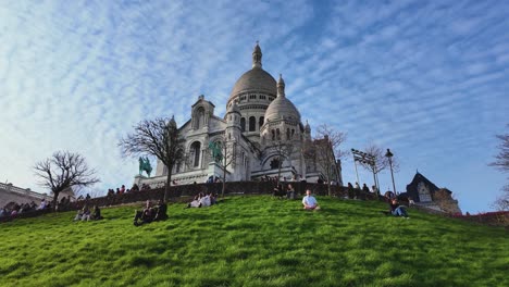 Die-Kamera-Bewegt-Sich-Zur-Seite-Der-Berühmten-Basilika-Sacré-Cœur-Im-Oberen-Teil-Der-Region-Montmartre-–-Menschen-Sitzen-Im-Gras-Rund-Um-Die-Kathedrale-Und-Genießen-Den-Sonnenuntergang-Am-Abend