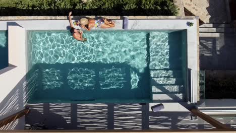 Brunette-woman-in-swimming-costume-lying-at-the-edge-of-the-pool-at-Cayo-Resort-in-Greece-Crete---man-next-to-her-drinks-a-cocktail-and-talks-to-her---drone-flies-over-them-in-bird's-eye-view