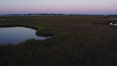 Swamp-wetlands-with-open-pools-and-tall-grass-reeds-and-homes-on-horizon-at-sunset,-Chincoteague-Island-Virginia,-slow-motion