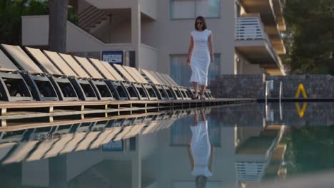 Attractive-woman-in-long-dress-walk-by-beach-hotel-pool-and-line-of-deck-chairs