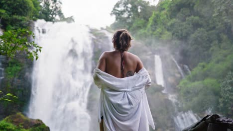 Woman-spreading-her-arms-and-letting-a-white-shirt-flow-in-the-wind,-standing-in-front-of-a-powerful-waterfall