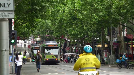 Eine-Urbane-Straßenszene-Zeigt-Straßenbahnen,-Die-Entlang-Der-Von-Bäumen-Gesäumten-Swanston-Street-Fahren,-Während-Essenslieferanten-Auf-Dem-Fahrradweg-In-Melbournes-Geschäftigem-Stadtzentrum-Unterwegs-Sind