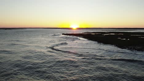 Drone-orbit-right-with-yellow-ball-of-sun-setting-on-distant-horizon-with-ocean-waves,-Chincoteague-Island-Virginia,-slow-motion