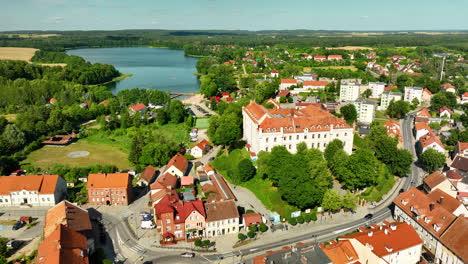 Vista-Aérea-De-La-Ciudad-De-Ryn,-Polonia,-Destacando-Edificios-Con-Techos-Rojos,-Exuberante-Vegetación-Y-Un-Sereno-Lago-Al-Fondo,-Capturando-Una-Mezcla-De-Belleza-Natural-Y-Encanto-Urbano.