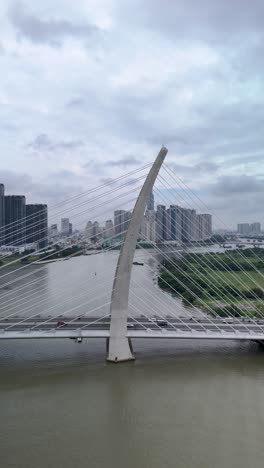 Traffic-crossing-Saigon-River,-Ho-Chi-Minh-City-overf-Ba-Son-suspension-Bridge-with-landmark-building-in-vertical-format-as-boat-passes-under