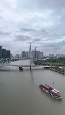 Container-boats-on-Saigon-River,-Ho-Chi-Minh-City,-with-aerial-view-of-Ba-Son-suspension-Bridge-with-landmark-building-in-vertical-format-as-boat-passes-under