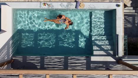 Lovely-couple-enjoying-the-pool-at-Cayo-Resort-in-Greece,-Crete---man-holds-brunette-woman-in-swimming-costume-in-water---drone-flies-in-bird's-eye-view-over-them