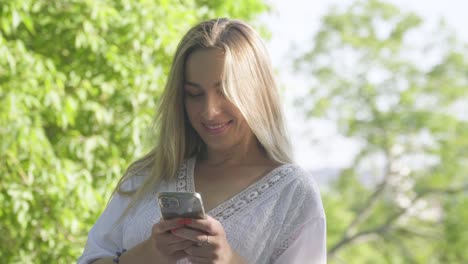 A-pretty,-happy-blonde-woman-in-a-white-dress-enjoying-a-happy-texting-conversation-in-the-summer-park