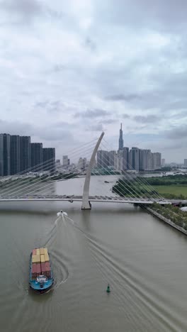 Container-boats-on-Saigon-River,-Ho-Chi-Minh-City,-with-aerial-view-of-Ba-Son-suspension-Bridge-with-landmark-building-in-vertical-format-as-boat-pass-along-river