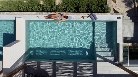 Mujer-Morena-En-Traje-De-Baño-Tumbada-En-El-Borde-De-Una-Piscina-En-Cayo-Resort-En-Grecia,-Creta---Tiene-Un-Cóctel-En-La-Mano-Y-Se-Acuesta---El-Dron-Vuela-Hacia-Arriba