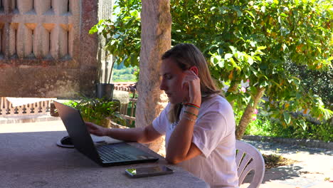 Young-blonde-woman-concentrating-on-teleworking-with-laptop-on-garden-table