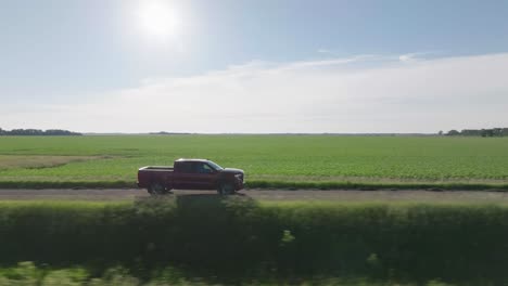 Aerial-drone-following-shot-over-a-vehicle-riding-on-route-through-green-farmlands