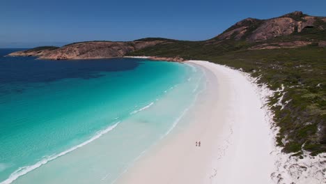 4k-Drone-Video-of-a-couple-walking-along-the-white-sand-beach-next-to-the-crystal-clear-blue-ocean-at-Thistle-Cove,-Esperance-WA