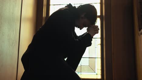 Young-man-in-old-church-building-in-front-of-stained-glass-window-kneeling,-praying,-worshipping