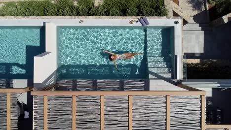 Brunette-woman-in-swimming-costume-enjoying-the-pool-at-CayoResort-in-Greece-Crete---she-swims-on-her-back-and-enjoys-the-water---drone-flies-in-bird's-eye-view