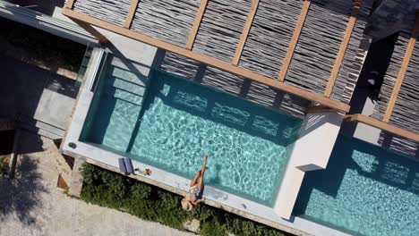 Brunette-woman-in-swimming-costume-is-sitting-at-the-edge-of-a-pool-in-Cayo-Resort-in-Greece-Crete---she-has-her-feet-in-the-water---drone-flies-down-and-turns-around