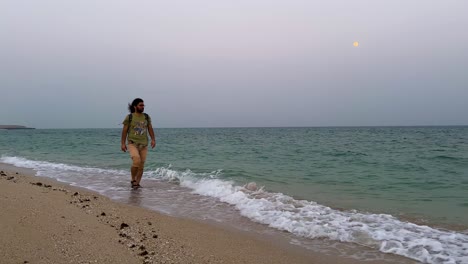 El-Hombre-Está-Caminando-A-Lo-Largo-De-La-Playa,-La-Línea-Del-Mar,-Usa-Una-Camisa-Verde-Oliva-Y-Un-Pantalón-Corto-En-La-Temporada-De-Verano,-Temprano-En-La-Mañana,-Cabello-Rizado,-Cabello-Largo,-Viaje-Masculino,-Pueblo-Rural,-Campo,-Crepúsculo,-Irán,-Paisaje-Natural.