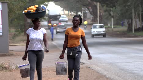 Mujeres-Africanas-Caminando-Cerca-De-Una-Calle-Muy-Transitada,-Una-Con-Su-Cesta-De-Compras-En-La-Cabeza