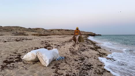 Mann-Im-Orangefarbenen-T-Shirt-Trägt-Strandmüll-Und-Zwei-Große-Müllsäcke-An-Malerischen-Sandstränden-In-Den-Emiraten.-Der-Panoramablick-Auf-Die-Tourismuslandschaft-Fördert-Den-Umweltschutz-In-Der-Kultur-Saudi-Arabiens