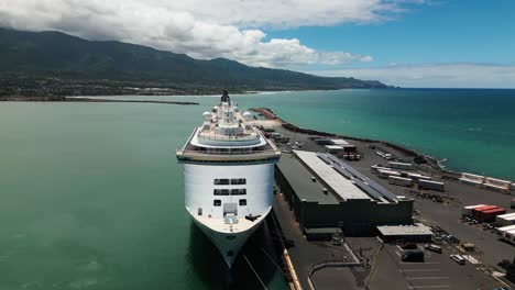 Drone-flies-toward-large-cruise-ship-docked-in-Maui-port-with-mountains-in-background