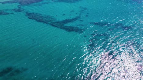 Drone-flies-over-shallow-ocean-floor-with-coral-and-rock-formations,-sunlight-reflecting-on-clear-blue-water