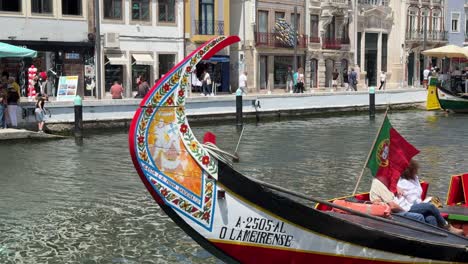 Point-of-view-of-the-colorful-and-traditional-Moliceiro-Boats-and-Portuguese-flag-waving-on-the-breeze-in-Aveiro,-Portugal