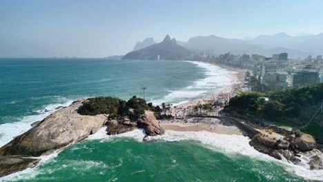 Atemberaubende-Drohnenaufnahmen-Von-Rio-De-Janeiros-Berühmtem-Strand-Von-Ipanema-An-Einem-Strahlend-Sonnigen-Tag,-Mit-Wunderschönen-Meereswellen,-Majestätischen-Bergen-Und-Der-Skyline-Der-Stadt-Am-Horizont