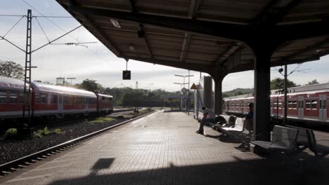 Early-morning-at-a-German-train-station,-light-casts-shadows-on-the-platform,-passengers-wait-as-train-approaches