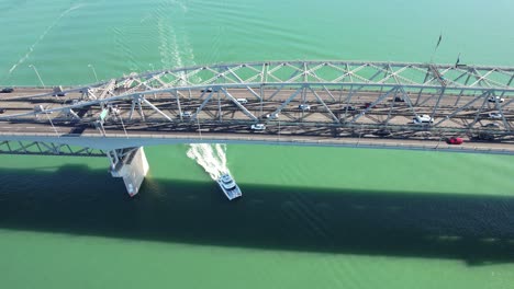 Catamaran-Boat-Cruising-Underneath-Auckland-Harbour-Bridge-With-Vehicles-Driving-Through-Box-Truss-Structure-In-New-Zealand