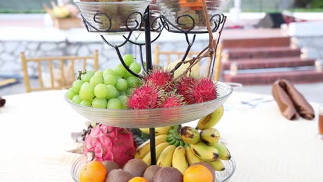 Centro-De-Mesa-De-Recepción-De-Boda-Con-Una-Bandeja-Escalonada-Que-Contiene-Uvas,-Plátanos,-Kiwi,-Rambután,-Pitahaya-Roja,-Lichi-Y-Naranjas,-Plataforma-Rodante-Lenta-De-Mano-Hacia-Adelante